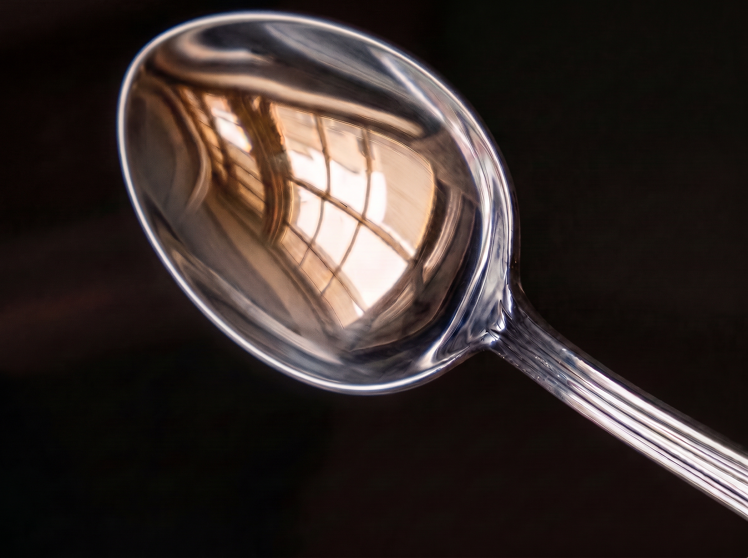 Un cucchiaio su un fondo scuro riflette la luce calda di una finestra, con i vetri incurvati nel metallo